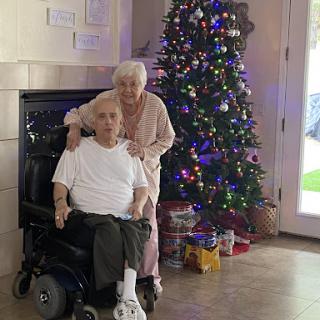 An elderly couple posing together in front of a beautifully decorated Christmas tree.