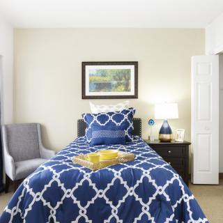 A cozy bedroom featuring a single bed with blue patterned bedding, an armchair, and a lamp on the nightstand.
