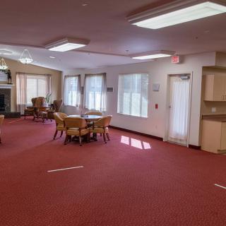 A spacious room with red carpet, featuring several tables and chairs, large windows, and overhead lighting.