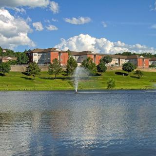 A serene view of a lake with a fountain in front of residential buildings and lush greenery under a bright blue sky.