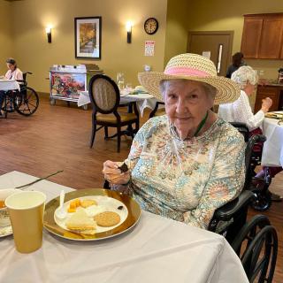 An elderly woman wearing a hat, sitting in a wheelchair and enjoying a meal in a dining area.