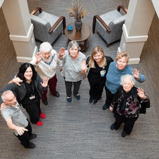 A cheerful group of seniors gathers together in a warm and inviting indoor space, happily waving at the camera.