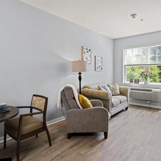 A cozy living room with a sofa and armchair, decorated in neutral tones and brightened by a window.