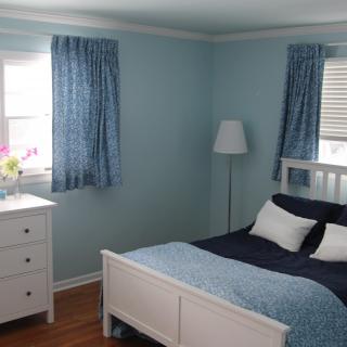 A cozy bedroom with blue walls and matching curtains, featuring a bed, dresser, lamp, and a vase of flowers.