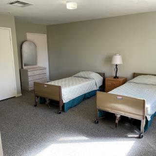 A minimalist bedroom with two hospital-style beds, a lamp, and a dresser against grey walls.