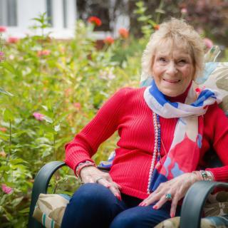 An elderly woman sitting comfortably in a garden, wearing a red sweater and colorful scarf.