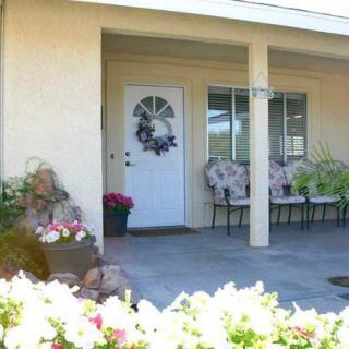 A cozy porch with a white door, windows, and patio chairs surrounded by flowers.