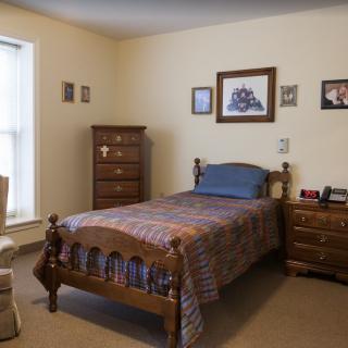 A cozy bedroom featuring a single wooden bed with a plaid comforter, accompanied by matching wooden furniture including a dresser and nightstand. The room is decorated with framed pictures on the walls and receives natural light from a large window.