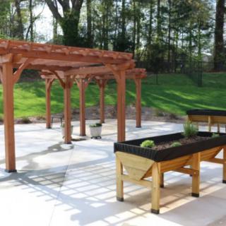 A patio area featuring a wooden pergola and raised garden beds, surrounded by greenery.