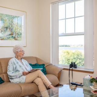 An elderly woman sits on a sofa in a well-lit living room, gazing out the window with binoculars nearby.