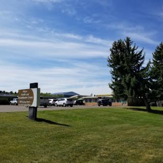 A sunny day view of a building with parked cars, surrounded by trees and open grass.