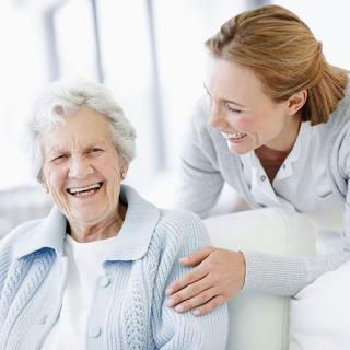 An elderly woman smiles warmly as her caregiver interacts with her supportively.