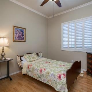 A cozy bedroom with a single bed and floral bedding, featuring a lamp on a nightstand, a piece of art on the wall, window blinds, and a ceiling fan.