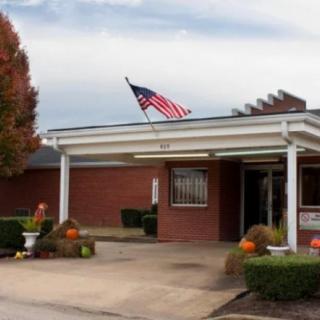 A brick building with an American flag flying above its entrance, surrounded by autumn trees and a parking area.