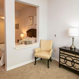 A well-lit bedroom with a plush chair, ornate dresser, and table lamp.