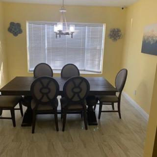A cozy dining room with a dark wooden table and six chairs, featuring a chandelier and large window with blinds against yellow walls.