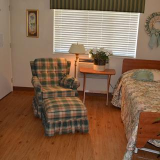 A cozy bedroom with a plaid upholstered chair and matching ottoman, a wooden bed, side table with a lamp, and an indoor plant by the window.