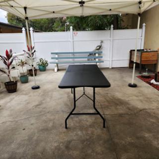 A black folding table set up under a tent in an outdoor patio area, surrounded by potted plants and a white fence.