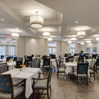A spacious dining room with multiple tables and chairs, set with white tablecloths, under soft lighting.