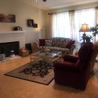 A cozy living room with a sofa and armchair arranged around a fireplace, featuring a decorative rug, a lamp, and large windows with curtains.