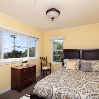 A cozy bedroom with yellow walls, featuring a bed, dresser, and large windows allowing natural light.