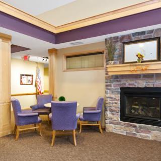 A cozy seating area with purple chairs arranged around a table, featuring a warm fireplace and carpeted flooring.