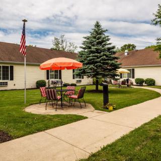 A cozy outdoor patio area with a table, chairs, and an umbrella set on a lush lawn, featuring a pathway leading through the garden with an American flag in the background.