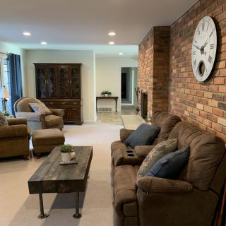 Cozy living room with a brick accent wall, featuring comfortable sofas and armchairs, a rustic coffee table, and a large wall clock.