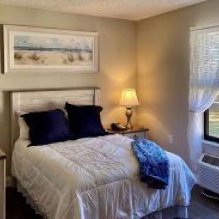 A cozy bedroom with a neatly made bed, featuring dark pillows and a blue throw. A lamp sits on the bedside table next to artwork above the headboard.