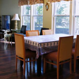 A dining room with a table covered by a cloth, surrounded by chairs and illuminated by natural light through large windows.