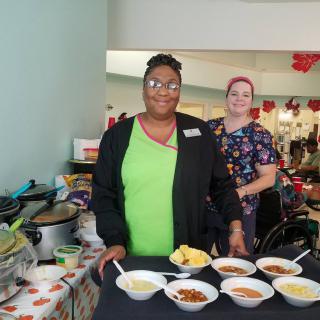 Two people serving food at an event with various dishes displayed.