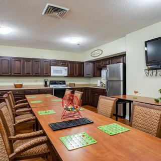 A kitchen area with a long table surrounded by chairs, featuring cabinets, a microwave, refrigerator, and a wall-mounted television.