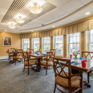A cozy dining room with neatly arranged tables and chairs, large windows allowing natural light in, and warm lighting fixtures.