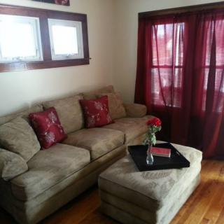 A cozy living room with a beige sectional sofa adorned with red cushions, accompanied by a matching ottoman and a decorative centerpiece on top. The room features wooden flooring, large windows with red curtains, and natural light filtering in.