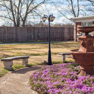 A serene garden setting with a fountain, purple flowers, benches, and a lamp post.