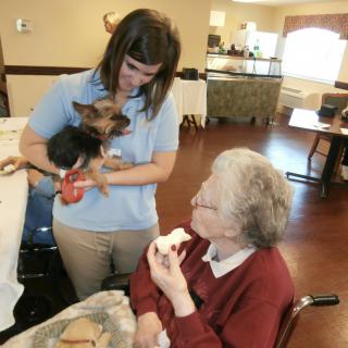 A nurse holds a small dog while interacting with an elderly woman in a care facility.