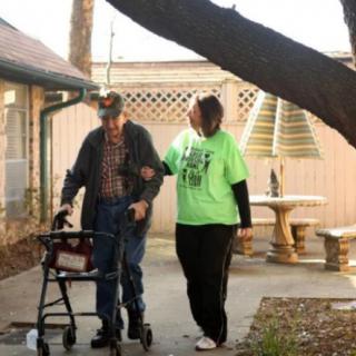 A person assists an elderly man using a walker outside.