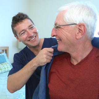 A caregiver assists an elderly man, both smiling in a positive interaction.