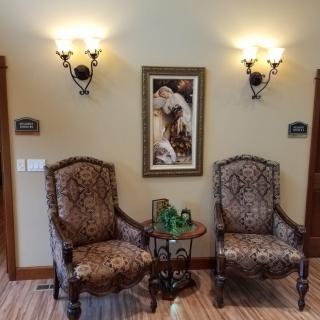 A cozy interior with two ornate chairs, a small side table, and framed wall art, illuminated by elegant sconces.