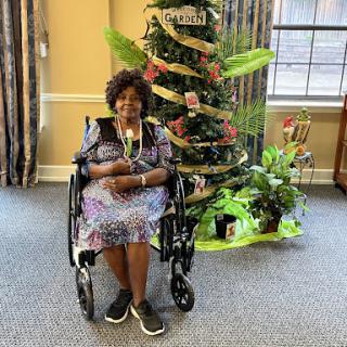 A cheerful woman in a wheelchair poses beside a beautifully decorated tree surrounded by greenery.
