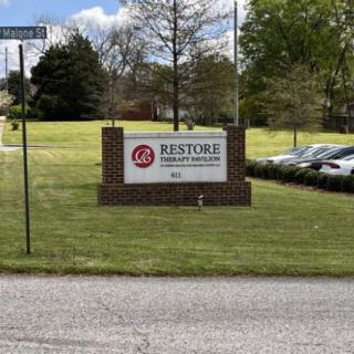 A business sign for Restore Place Counseling stands on a grass lawn next to a parking lot with several cars.