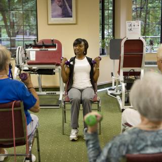 A fitness instructor leads a group of seniors in chair exercises using weights.