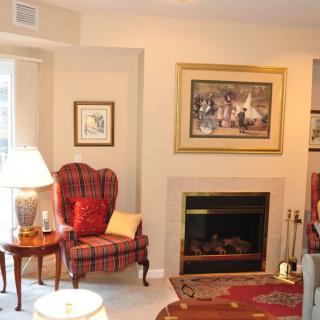 A cozy living room with a plaid armchair and fireplace, adorned with artwork and a lamp.