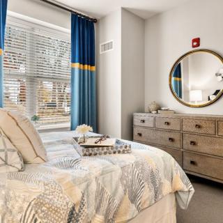 A cozy bedroom featuring a wooden dresser, round mirror, and a bed with patterned bedding, illuminated by natural light through the window.