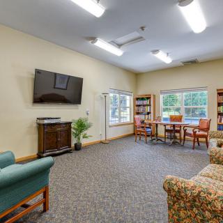 A cozy living room with a television, bookshelves, chairs, and large windows.