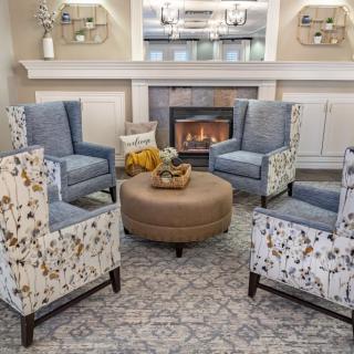 A cozy living room with patterned armchairs arranged around a central ottoman, set in front of a lit fireplace.