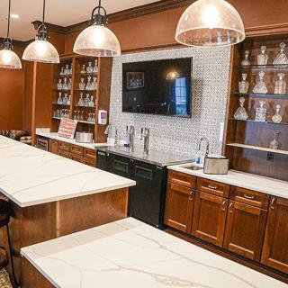 A modern kitchen interior featuring wood cabinets, elegant countertops, shelving units, pendant lights, and a large TV screen.