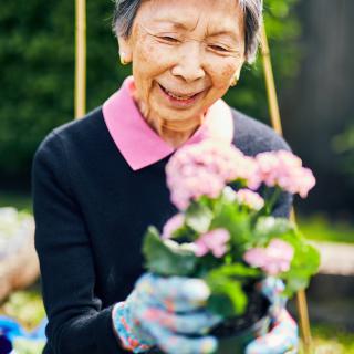 An elderly woman happily tending to her garden, holding a pot of pink flowers.