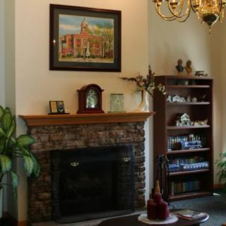 A cozy living room with a stone-faced fireplace, surrounded by decor, a clock on the mantel, a painting above, and a wooden shelf filled with ornaments.