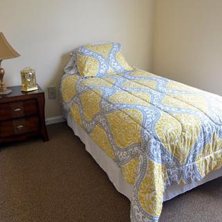 A cozy bedroom featuring a single bed with a yellow and blue quilt, next to a wooden nightstand with a lamp.